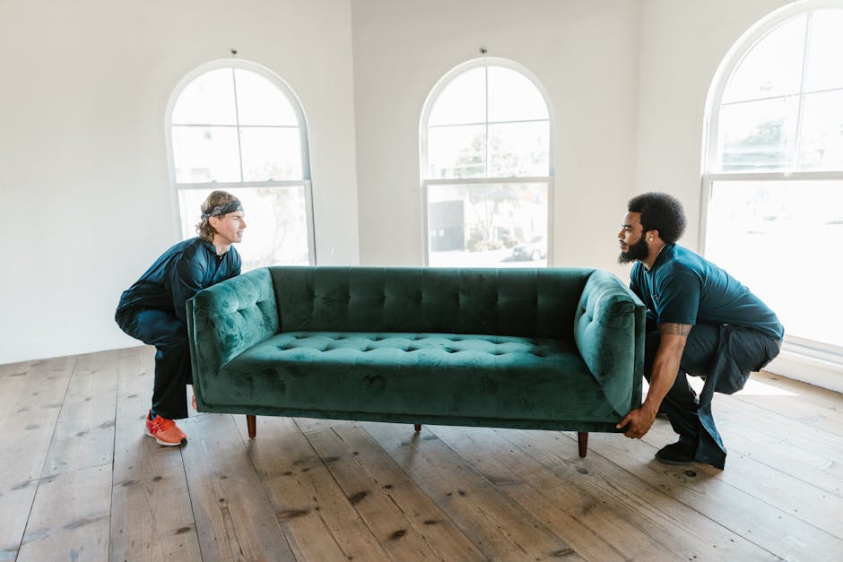 Two movers from Man with Van Leaves Green are engaged in the home relocation process, lifting a green upholstered sofa with a smooth fabric finish and wooden legs, positioning it through a clear doorw