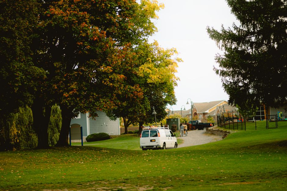 A white moving van parked on a residential street in Leaves Green, surrounded by green grass and mature trees with changing autumn leaves. The van is positioned near the driveway of a house, partially visible in the background, with its rear doors open as a man and a woman unload cardboard boxes and furniture wrapped in protective materials. The street is narrow, with outdoor lighting fixtures and a gentle slope leading from the van to the house, indicating a typical home relocation scenario. Towels, packing blankets, and plastic wrapping are visible, used to secure and protect items during the furniture transport and loading process, with some boxes placed on trolleys for easier movement. The scene reflects the logistics of packing and moving on a narrow street or tight drive, with visible signs of careful planning by Man with Van Leaves Green to facilitate efficient house removals.